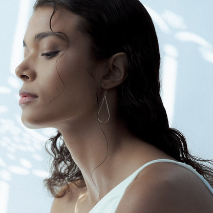Woman with closed eyes against a white wall in Greece, wearing an earring in silver with the shape of a Raindrop, by Aurore Havenne