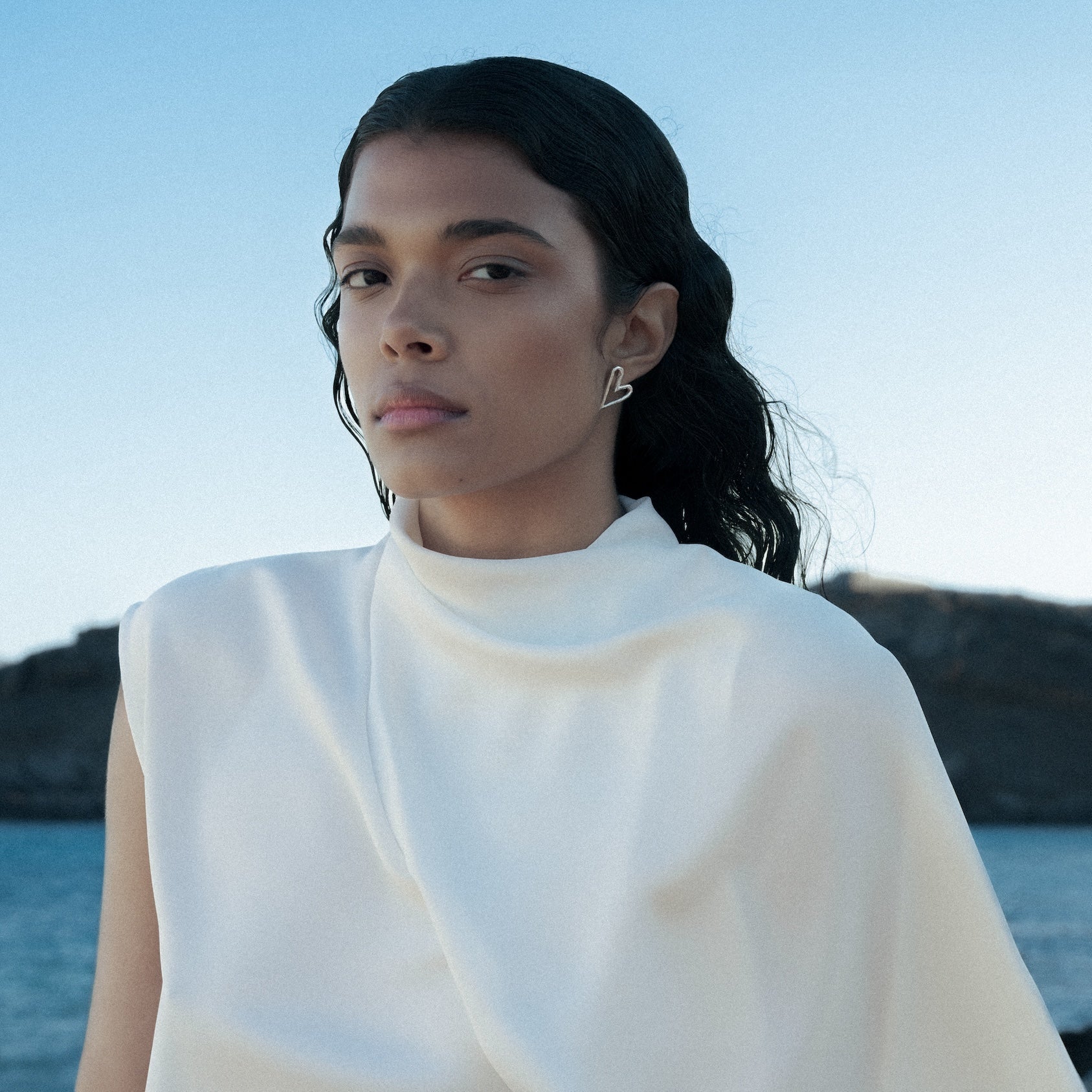 A woman with dark hair tied back wears a white draped sleeveless top and heart shaped earrings by Aurore Havenne, standing outdoors near a body of water with rocky mountains in the background.