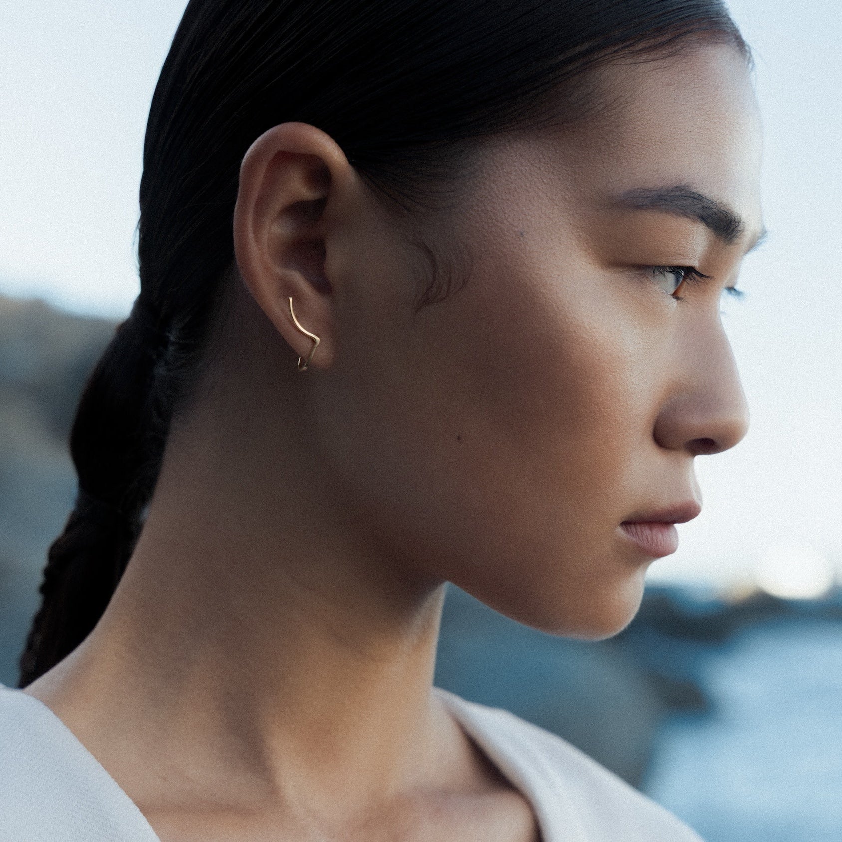A woman in profile wearing a delicate gold rhea earring by Aurore Havenne, photographed outdoors with a blurred coastal background.