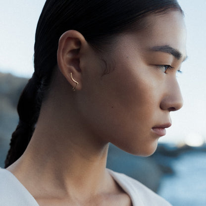 A woman in profile wearing a delicate gold rhea earring by Aurore Havenne, photographed outdoors with a blurred coastal background.