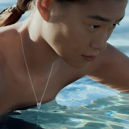 A close-up lifestyle shot of a model in shallow water wearing a delicate silver heart pendant necklace.