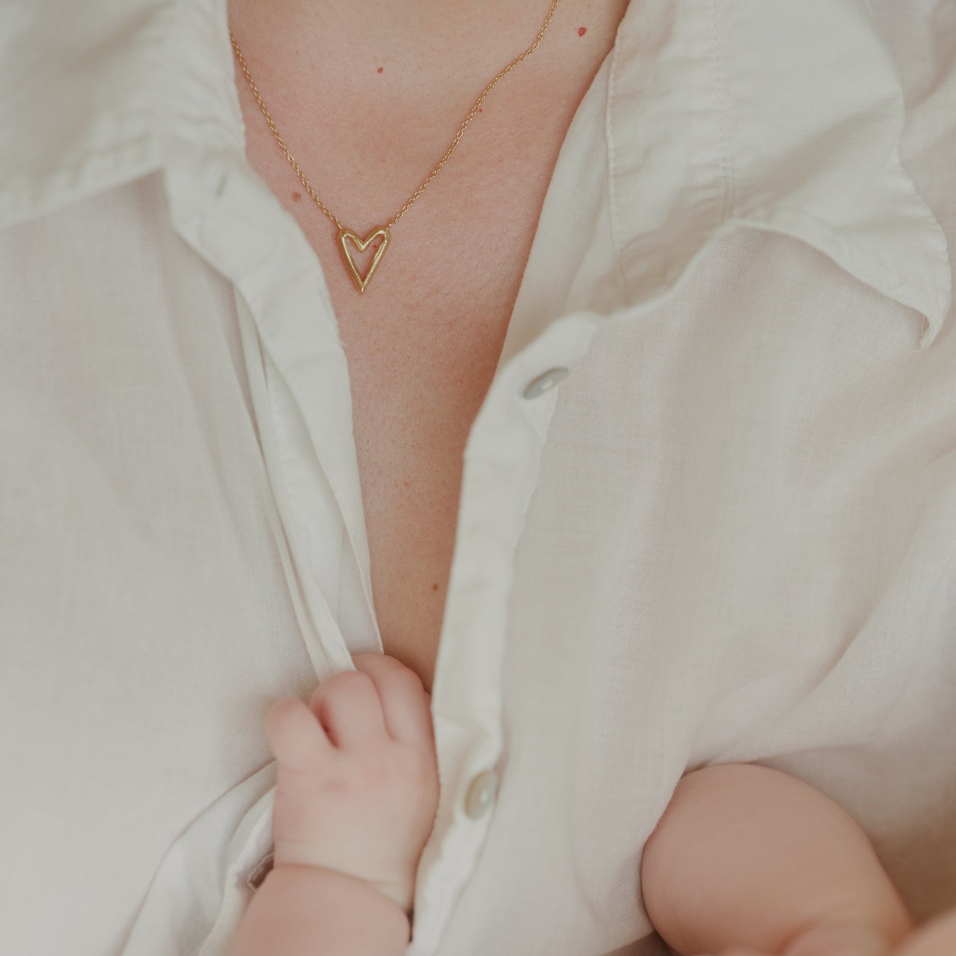 Close-up of a person wearing a heart-shaped necklace with a baby's hand holding onto their shirt.